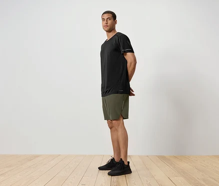 Seitenansicht eines Mannes in einem schwarzen T-Shirt, grünen Shorts und schwarzen Turnschuhen, der auf einem Holzboden vor einer weißen Wand steht.