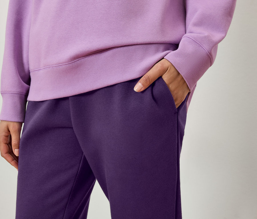 Detail einer Person in einem lila Pullover und dunkelvioletten Jogginghosen, eine Hand in der Tasche.