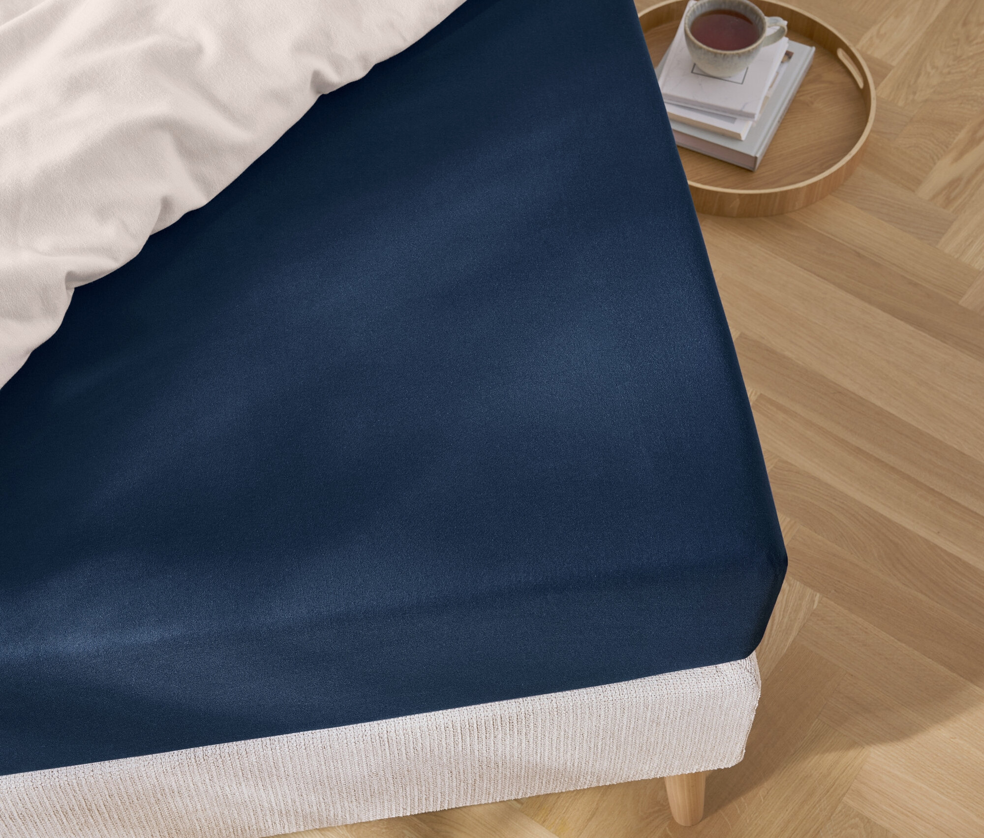 Detailansicht eines blauen Spannbettlakens auf einem Bett mit weisser Bettwäsche und einem Tablett mit Büchern und einer Tasse.