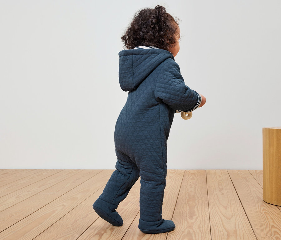 Kleinkind in blauem Steppoverall mit Kapuze geht auf Holzboden.