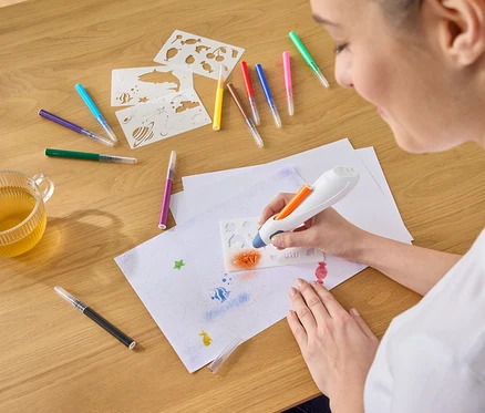 Frau sprüht mit einem Airbrush-Stift orange Farbe durch eine Schablone auf ein Blatt Papier, umgeben von Stiften, Schablonen und einer Tasse Tee.