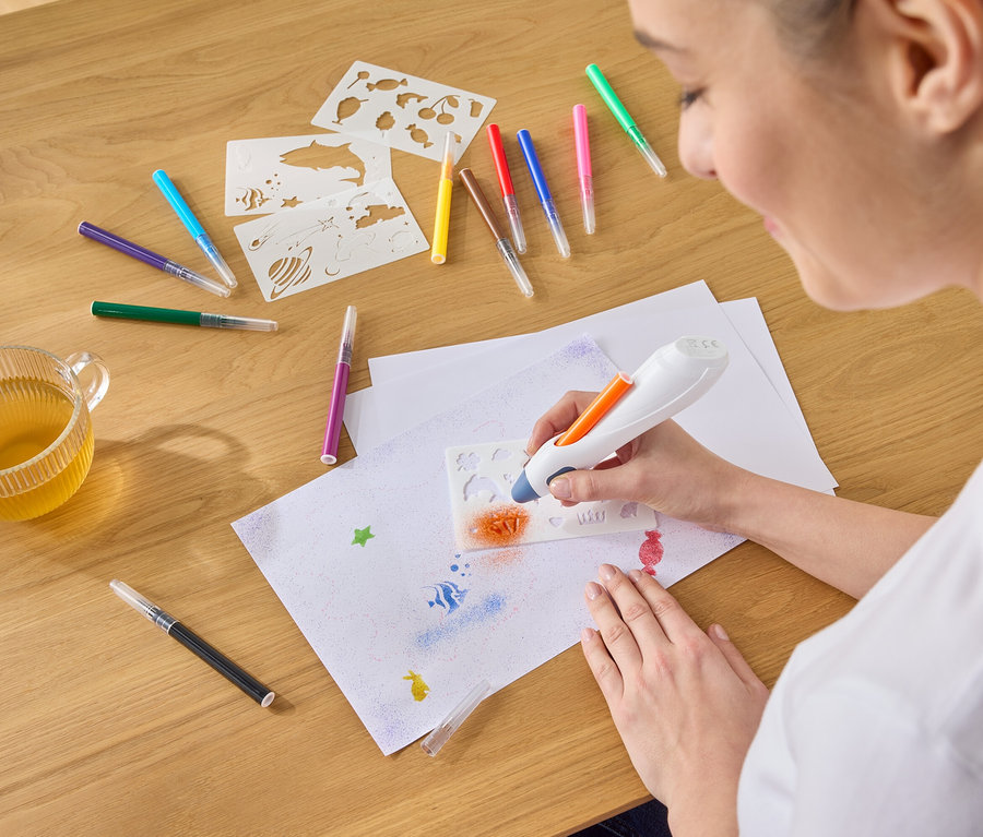 Frau sprüht mit einem Airbrush-Stift orange Farbe durch eine Schablone auf ein Blatt Papier, umgeben von Stiften, Schablonen und einer Tasse Tee.