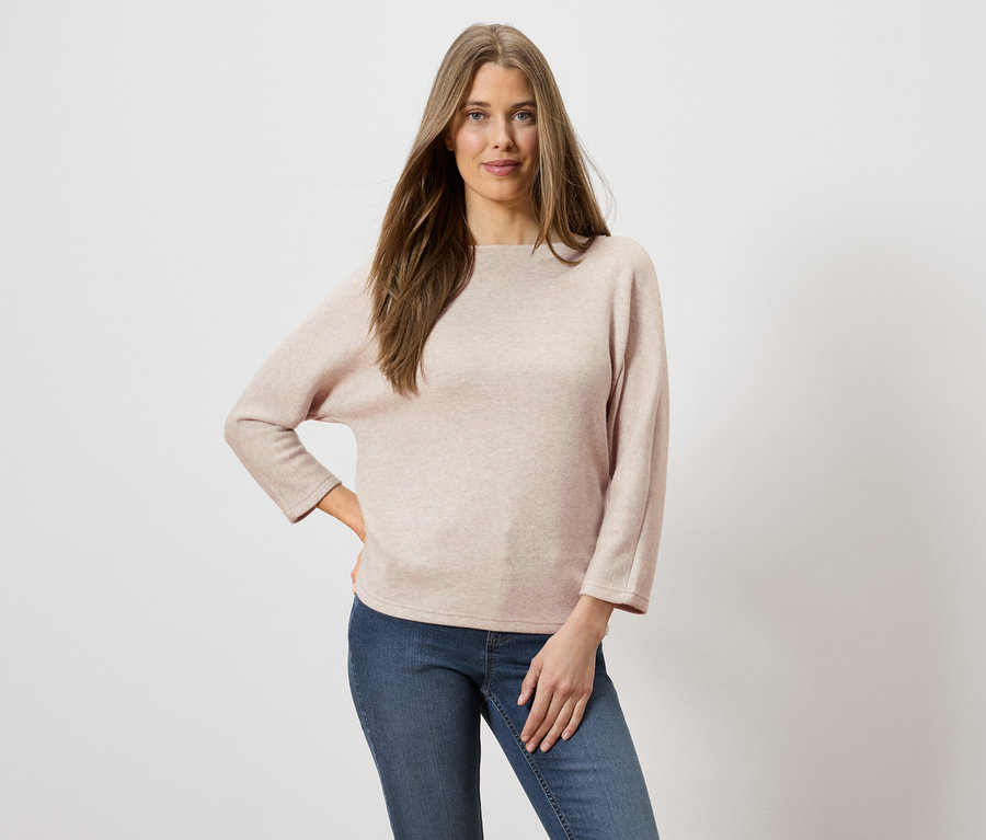Frau mit langen braunen Haaren, die einen hellrosa Pullover und Jeans trägt und mit einer Hand auf der Hüfte posiert.