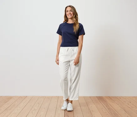 Frau mit langen, gewellten, braunen Haaren, die ein marineblaues T-Shirt, eine weiße Hose mit Kordelzug und weiße Turnschuhe trägt.