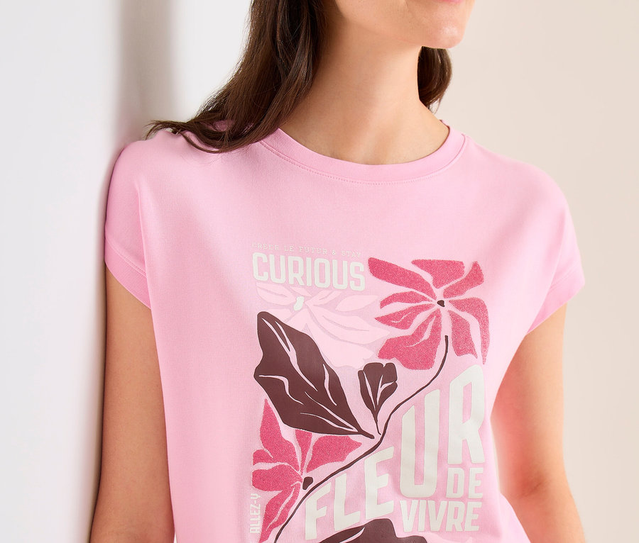 Frau mit beigem Cap und pinkem CECIL Placement-Shirt mit Blumenmuster und Schriftzug "FLEUR DE VIVRE" lehnt an einer Wand.