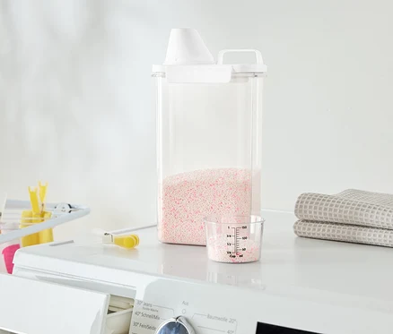 Auf einer Waschmaschine stehen ein durchsichtiger Behälter mit rosa Waschmittel, ein Messbecher und gefaltete Handtücher.