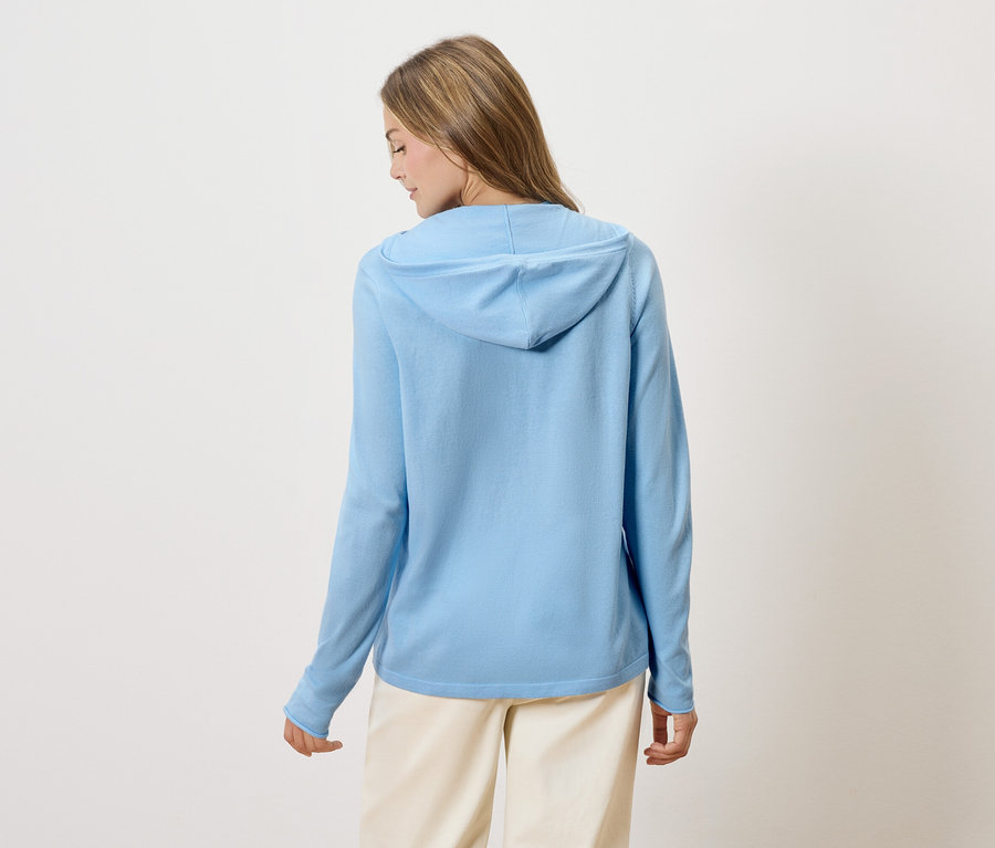 Rückansicht einer Frau mit blonden Haaren, die einen hellblauen Kapuzenpullover und eine beigefarbene Hose trägt.