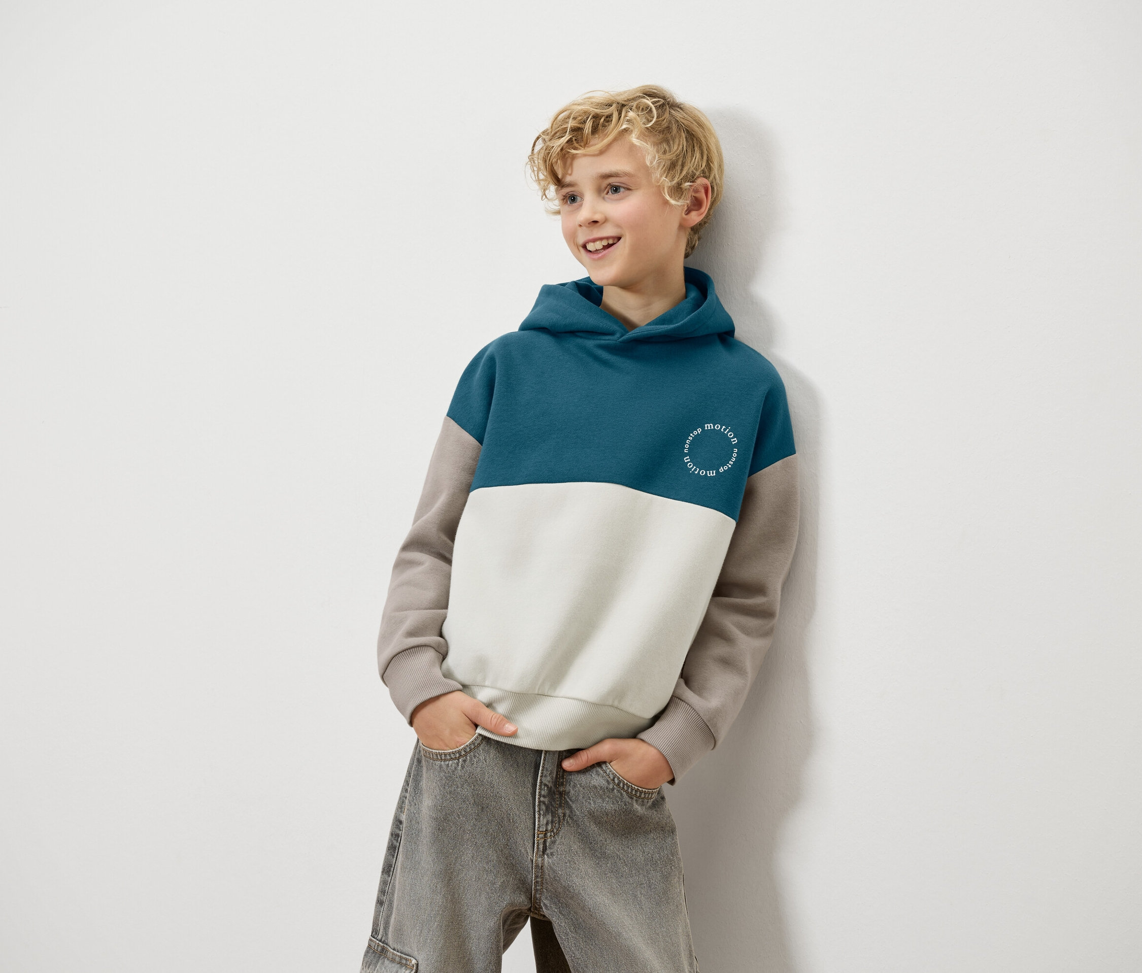 Ein blonder Junge lehnt an einer Wand und trägt einen blau-weiß-braunen Kapuzenpullover und graue Jeans.