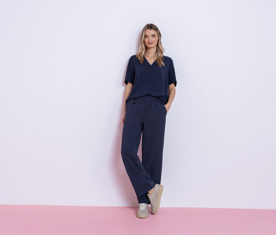 Frau steht vor einem rosa und weißen Hintergrund und trägt ein marineblaues Outfit, bestehend aus einem T-Shirt und einer Hose, sowie graue Turnschuhe.