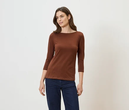 Frau mit braunem, langärmligem Shirt und Jeans vor hellem Hintergrund.