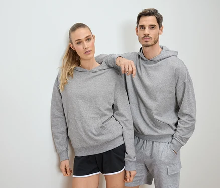 Frau und Mann posieren in grauen Hoodies und Shorts vor einer weißen Wand.