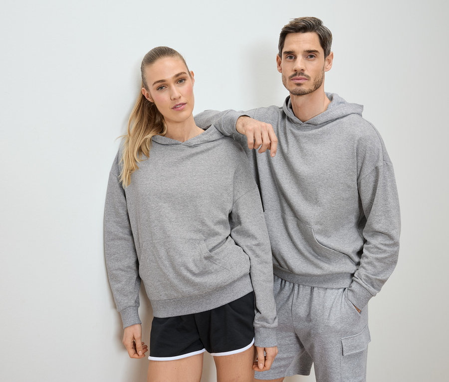 Frau und Mann posieren in grauen Hoodies und Shorts vor einer weißen Wand.