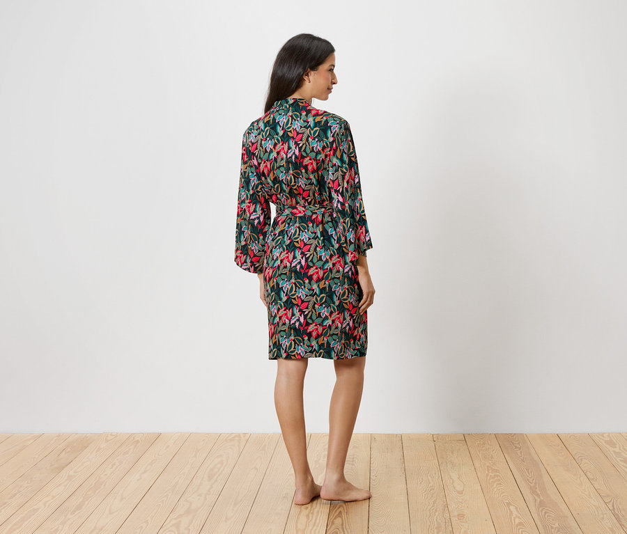 Frau von hinten, die einen floralen Morgenmantel mit Bindegürtel vor einer weißen Wand trägt.