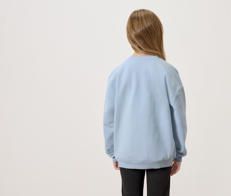 Rückansicht eines Kindes mit langen braunen Haaren, das einen hellblauen Pullover und schwarze Hosen trägt.