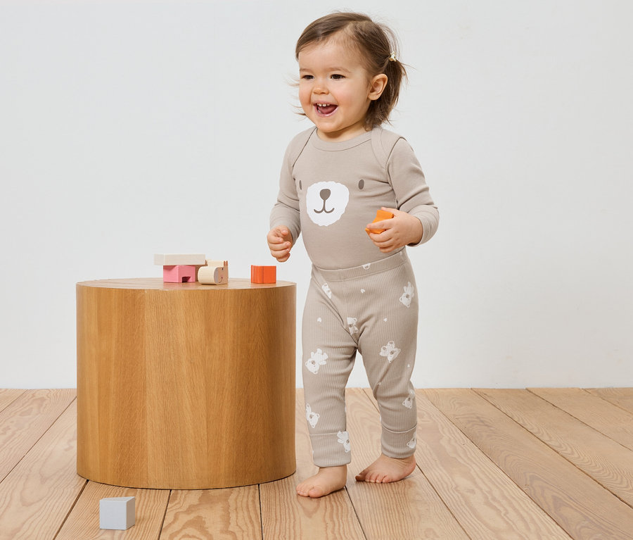 Ein lachendes Kleinkind in einem beigen Bären-Pyjama steht neben einem Holzhocker mit Spielzeug.