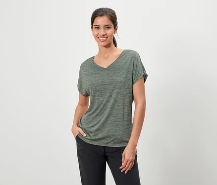 Frau mit dunklen Haaren, die ein meliertes olivgrünes T-Shirt und eine dunkle Hose trägt und mit einer Hand in der Tasche lächelt.