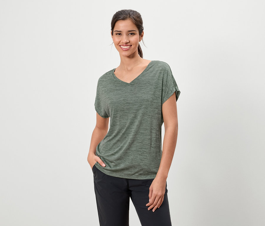 Frau mit dunklen Haaren, die ein meliertes olivgrünes T-Shirt und eine dunkle Hose trägt und mit einer Hand in der Tasche lächelt.