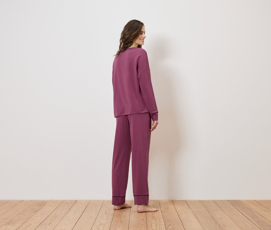 Frau von hinten in langärmligem, weinrotem Pyjama-Set auf Holzboden vor einer weißen Wand.