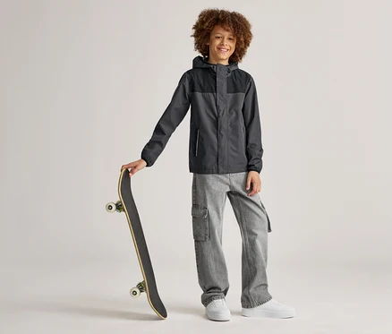 Ein lächelnder Junge mit lockigem Haar steht mit einem Skateboard in der Hand, trägt eine grau-schwarze Jacke, graue Jeans und weiße Schuhe.