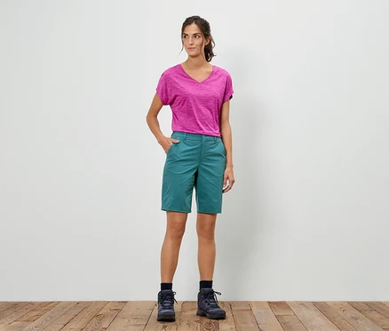 Frau steht mit der Hand in der Hosentasche, trägt ein pinkes T-Shirt, grüne Shorts und Wanderschuhe.