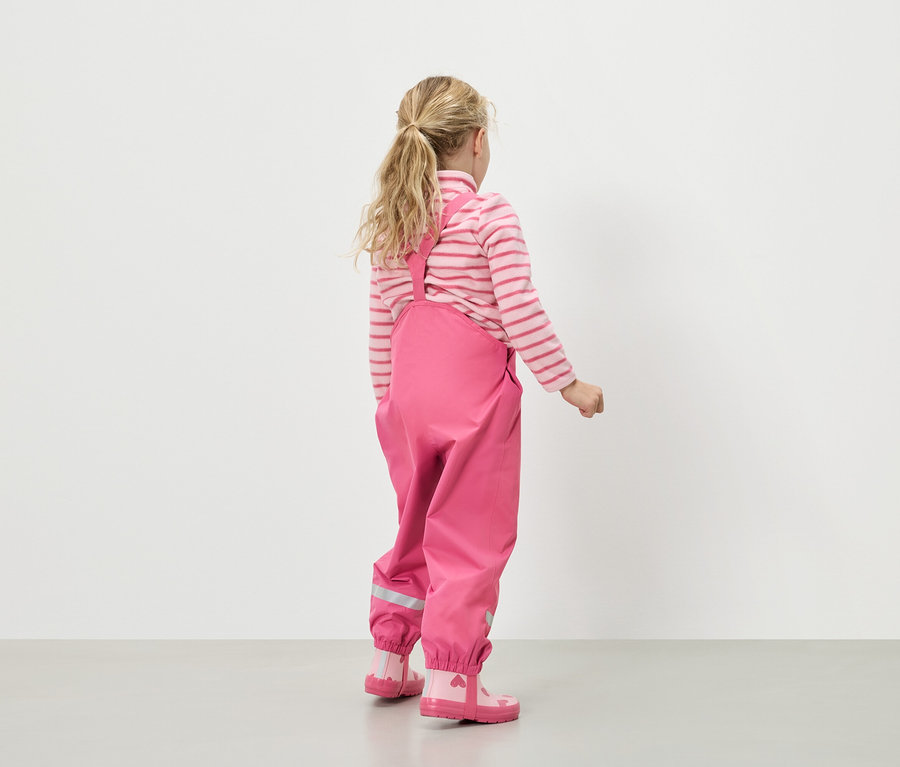 Rückansicht eines Mädchens mit blonden Haaren, das einen rosa-weiß gestreiften Pullover, eine rosa Regenhose mit Trägern und rosa Gummistiefel mit Herzen trägt.
