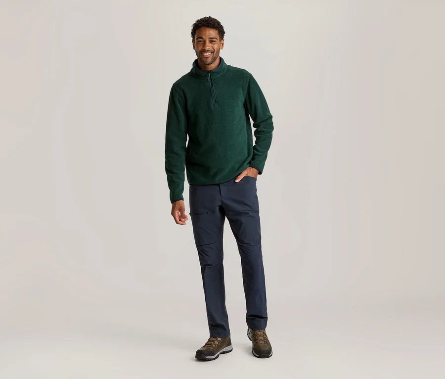 Ein lächelnder Mann steht und trägt einen dunkelgrünen Fleece-Pullover mit Reißverschluss, dunkelblaue Hosen und braune Wanderschuhe. Er hat die rechte Hand in der Hosentasche.