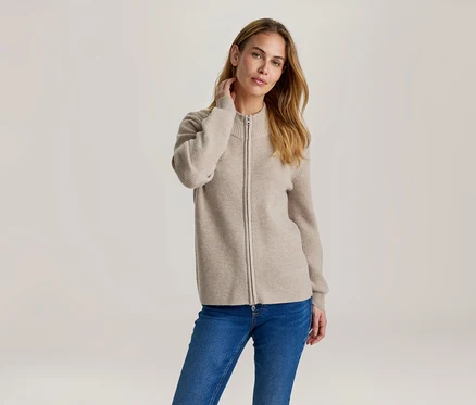 Frau mit langen, hellbraunen Haaren, die eine beigefarbene, gerippte Strickjacke mit Reißverschluss und blaue Jeans trägt. Sie berührt ihren Nacken.