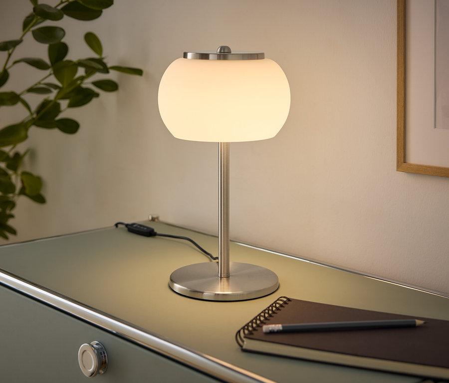 Eine leuchtende Tischlampe mit weißem Glaskugel-Schirm und gebürstetem Metallsockel steht auf einer hellgrünen Kommode. Daneben liegen ein Notizbuch und ein Bleistift. Links ist eine Pflanze, rechts ein gerahmtes Bild an der Wand.