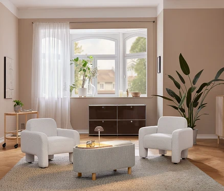Gemütliches Wohnzimmer mit zwei hellgrauen Sesseln und einem passenden Ottoman auf einem Teppich. Eine beige LED-Akku-Tischleuchte steht auf einem hellen Holzschrank neben einer großen Pflanze. Unter einem Erkerfenster befindet sich ein dunkelbraunes Aufbewahrungsmöbel.