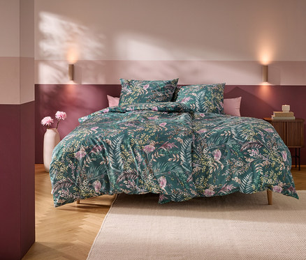 Ein Bett mit grünem Blumenmuster-Bettbezug und zwei Kissen in einem Schlafzimmer mit horizontal gestreiften rosa-burgunderroten Wänden. Zwei Wandleuchten beleuchten die Wand über dem Bett. Eine weiße Vase mit rosa Blumen steht links, ein Holznachttisch mit Büchern und einer Kerze rechts. Ein heller Teppich liegt auf dem Holzboden.