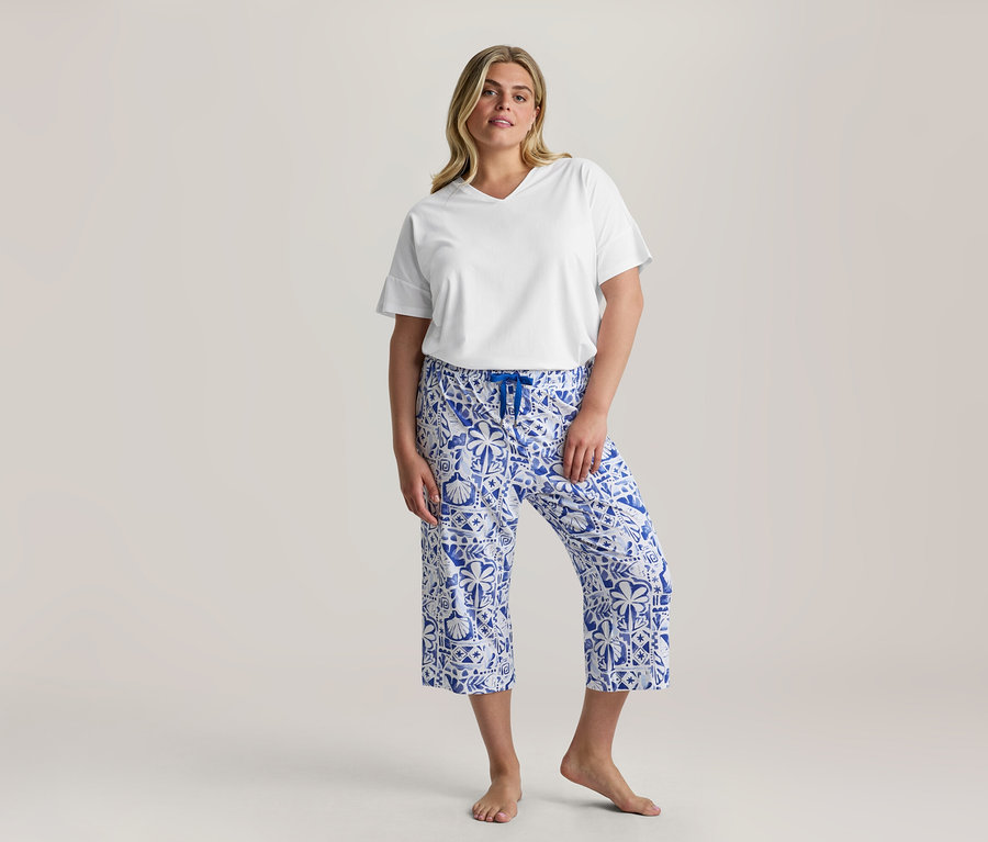 Frau steht, trägt ein weißes V-Ausschnitt-T-Shirt und eine blau-weiß gemusterte 3/4-Pyjama-Set Hose mit blauem Kordelzug, barfuß.