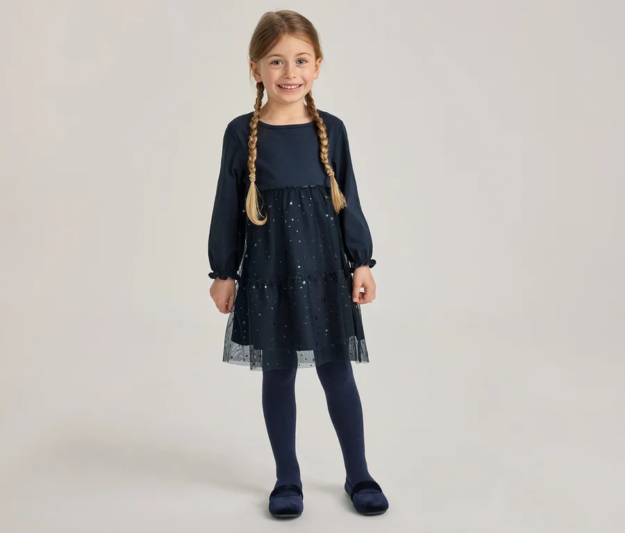 Lächelndes Mädchen in dunkelblauem, langärmeligem Kleid mit glitzerndem Tüllrock, dunkelblauer Strumpfhose und Samtschuhen. Sie trägt zwei Zöpfe.