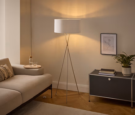 Ein gemütliches Wohnzimmer mit einem hellen Sofa, einer Stehlampe mit weißem Schirm und einem dunkelgrauen Schrank. Auf der Armlehne des Sofas steht ein Tablett mit einer Wasserflasche und einem Glas. Auf dem Schrank befinden sich eine Topfpflanze, Bücher und darüber hängt ein gerahmtes Line-Art-Bild.