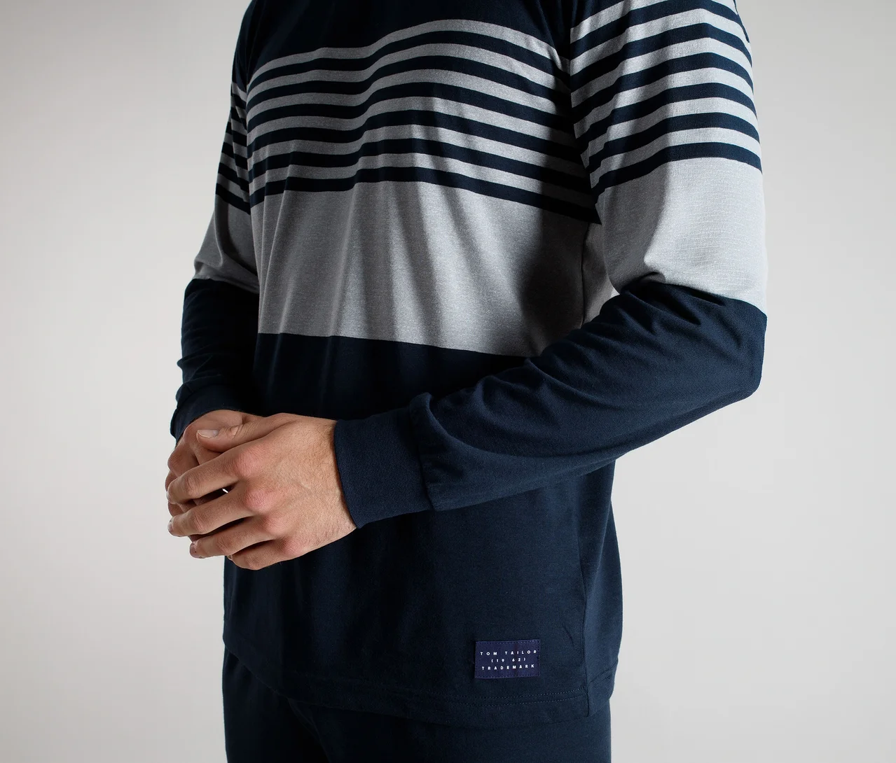 Mann trägt ein Langarmshirt mit horizontalen dunkelblauen und grauen Streifen oben, einem einfarbigen grauen Mittelteil und einem einfarbigen dunkelblauen Unterteil und Ärmeln. Seine Hände sind gefaltet.