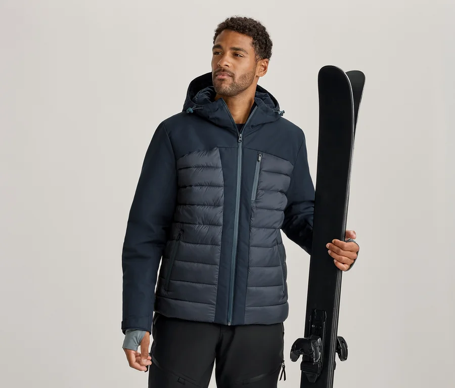 Mann in dunkelblauer und grauer Skijacke und schwarzer Skihose, der ein Paar schwarze Skier hält.