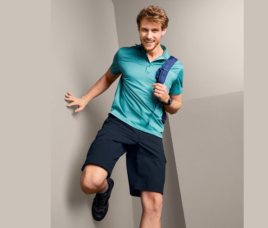 Mann lehnt an Wand, trägt Funktionsshorts, Poloshirt und Rucksack.