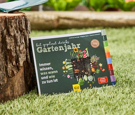 Das Buch »Gut geplant durchs Gartenjahr« liegt auf dem Rasen neben einem Baumstumpf, auf dem eine Gartenschaufel liegt.