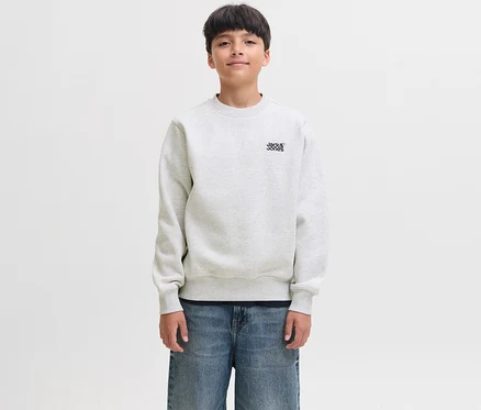 Ein junger Junge mit dunklen Haaren lächelt leicht und trägt einen weißen Jack&Jones Sweater Colton mit dem Markenlogo auf der Brust und blaue Jeans.