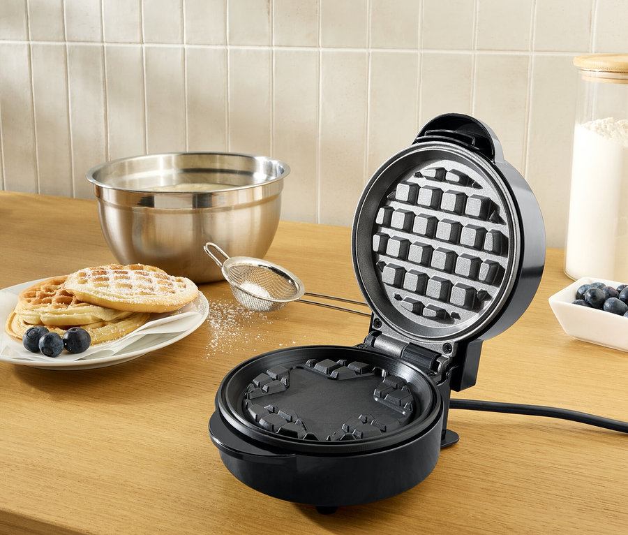 Ein offenes, schwarzes Waffeleisen auf einer hölzernen Arbeitsplatte, daneben ein Teller mit mit Puderzucker bestreuten Waffeln und Blaubeeren.