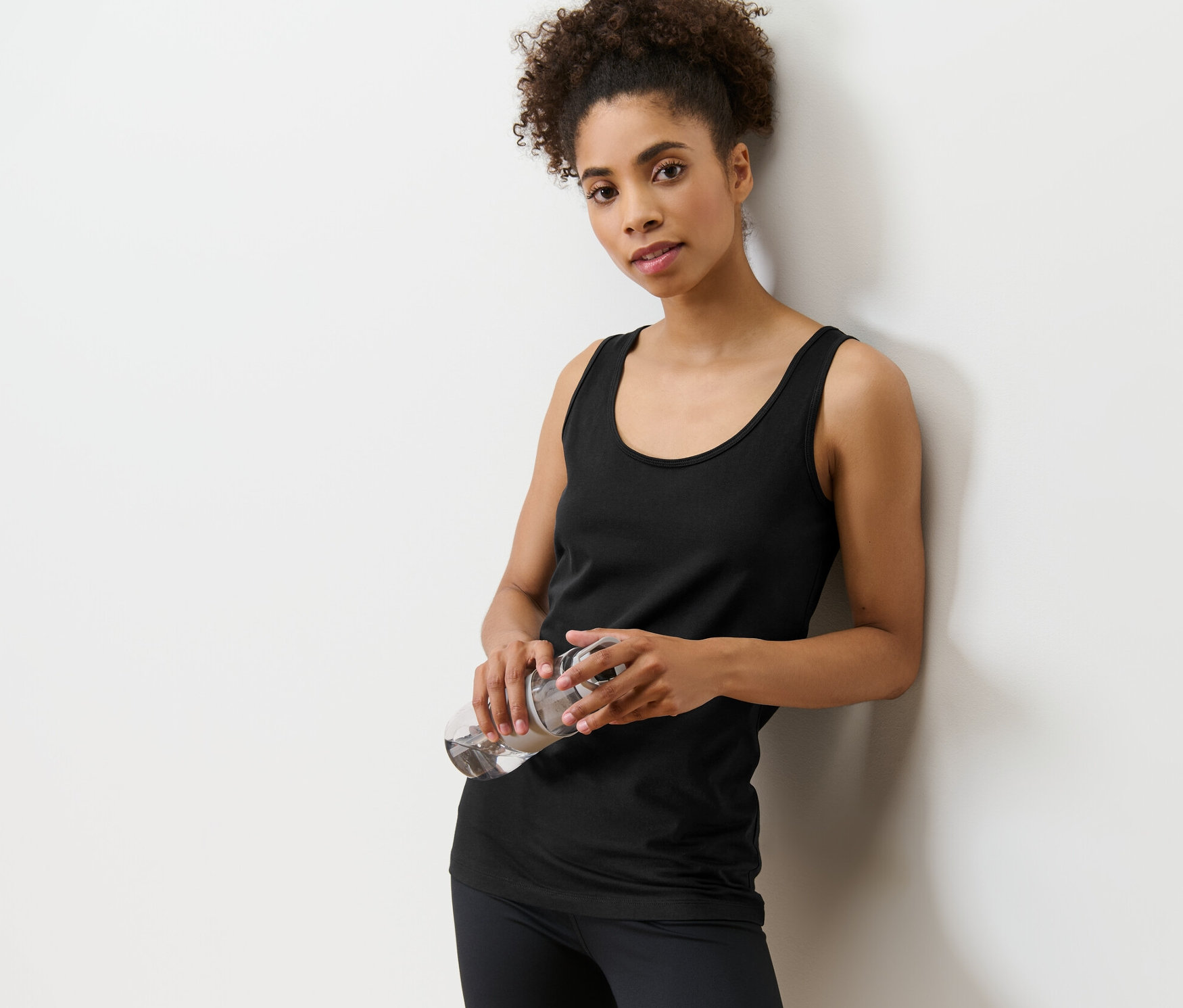 Frau, an eine Wand gelehnt, hält eine Wasserflasche. Sie trägt ein schwarzes Top aus einem 2er-Set.