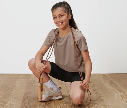 Mädchen in braunem Sportshirt aus dem 2 Kinder-Sportshirts Set mit schwarzer Hose hält ein Springseil.