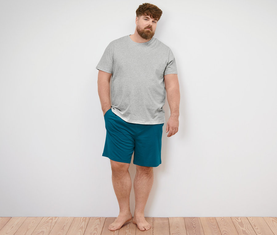 Mann steht barfuß an einer Wand, trägt ein graues T-Shirt mit Rundhalsausschnitt und blaue Pyjamashorts.