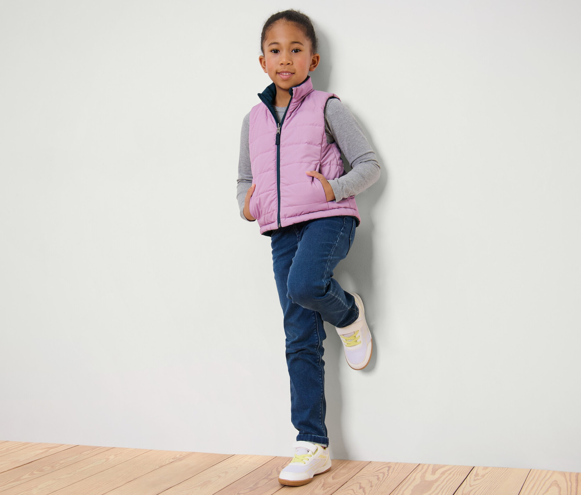 Mädchen, an eine Wand gelehnt, trägt eine Kinder-Wende-Steppweste, Jeans und Turnschuhe.