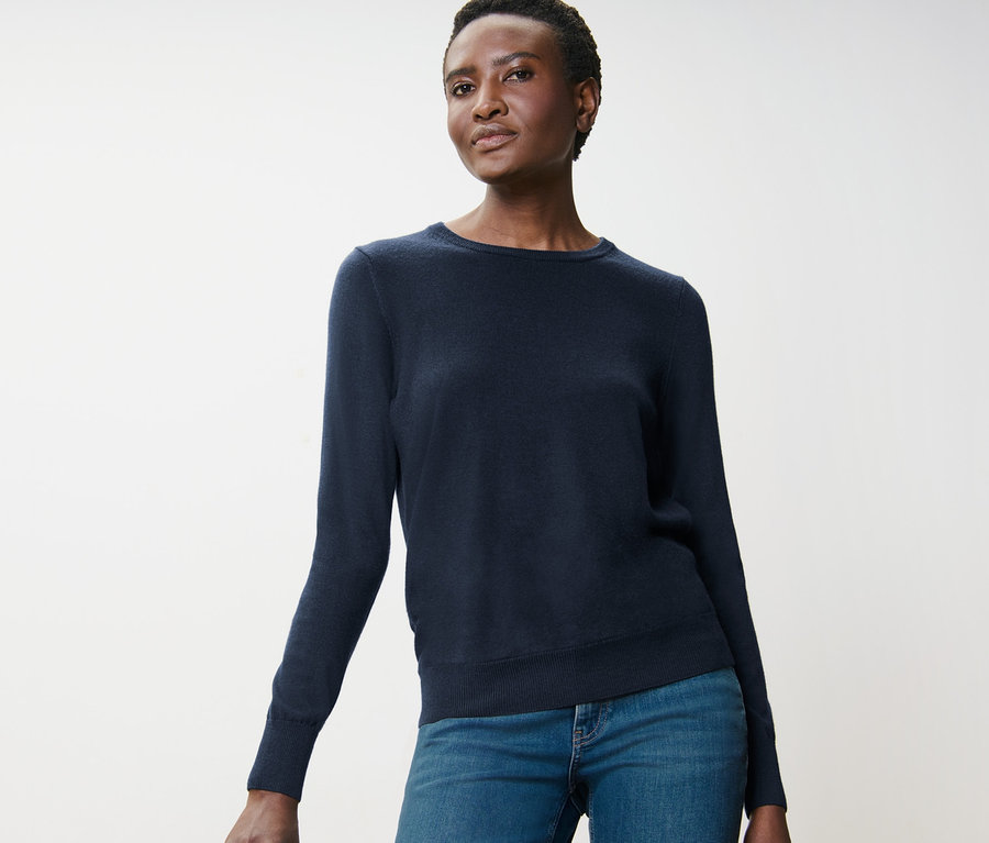 Eine Frau posiert in einem dunkelblauen Merino-Feinstrickpullover und Jeans neben einem Holzhocker.