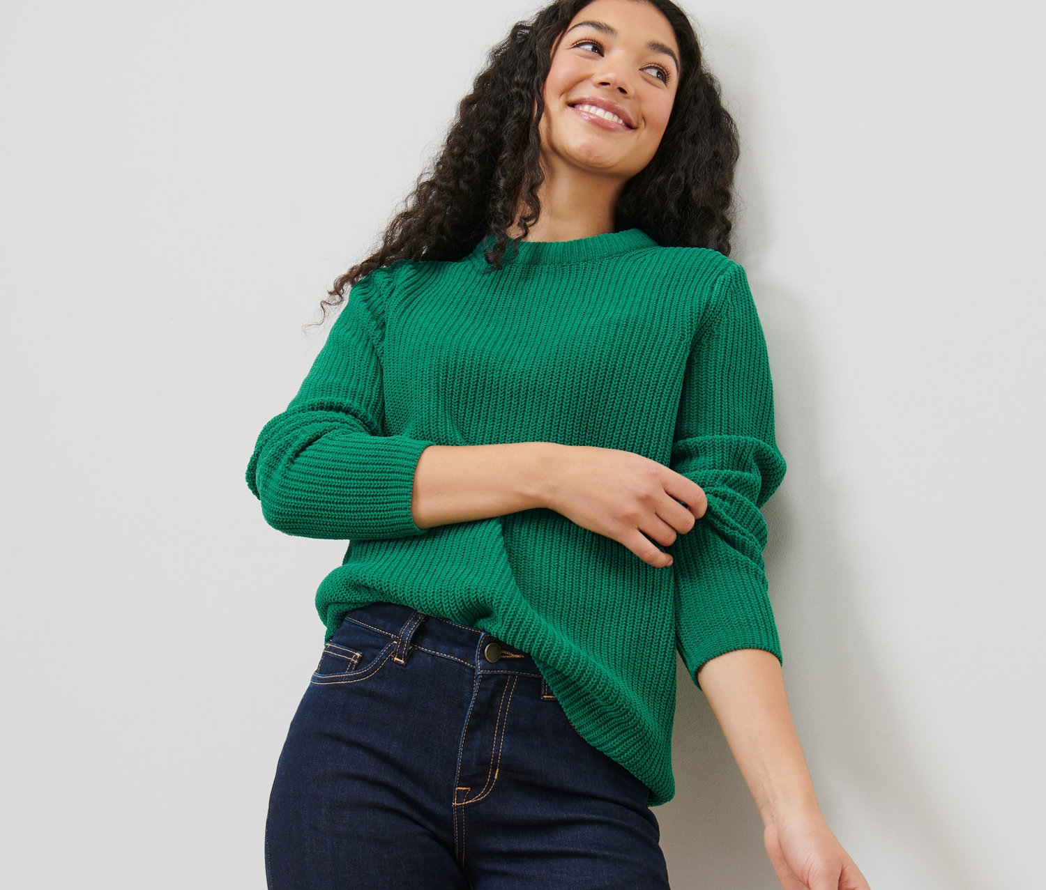 Eine Frau mit dunklen lockigen Haaren trägt einen grünen Grobstrickpullover und Jeans.