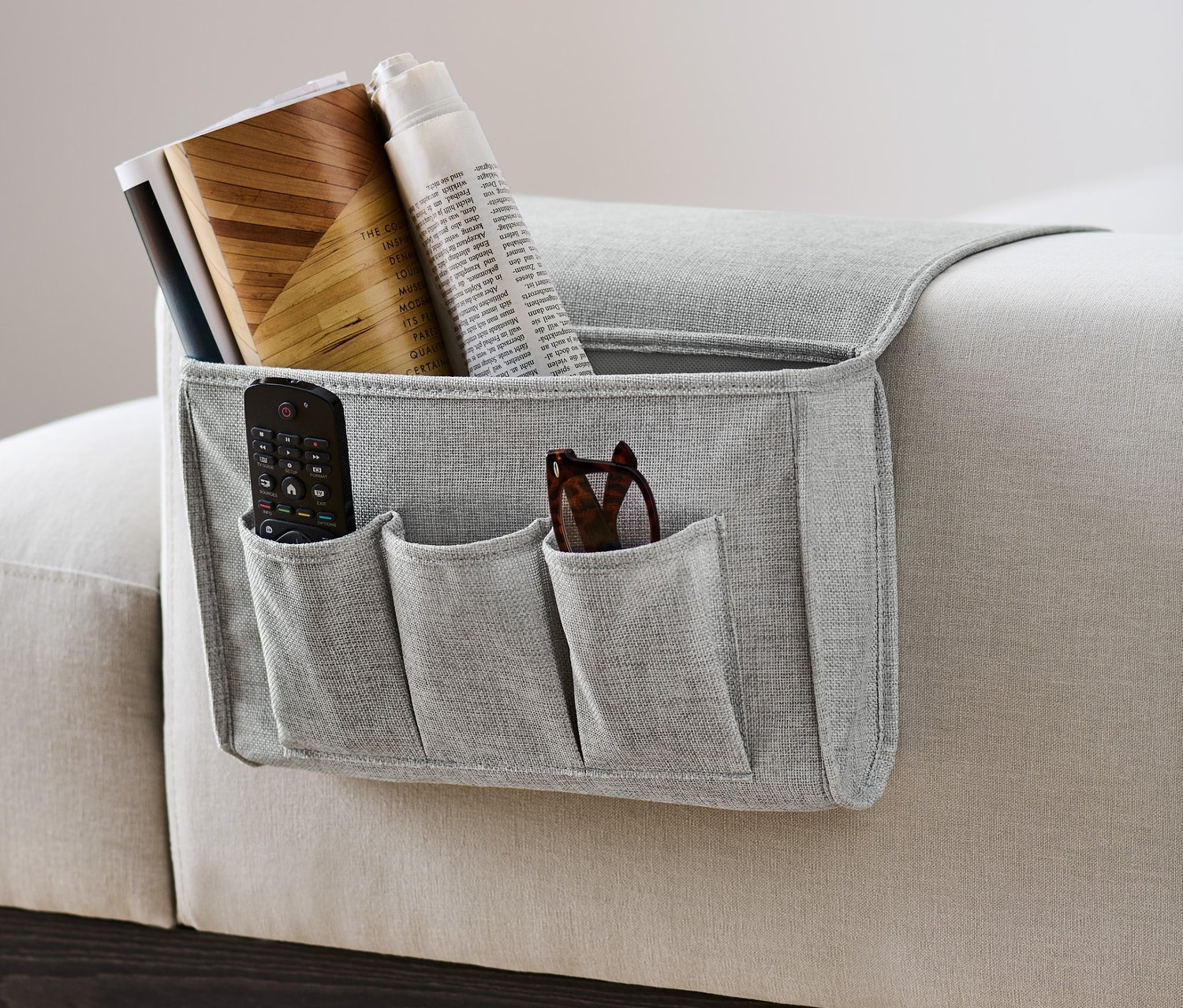 Grauer Bett-und-Couchorganizer hängt an der Seite eines Sofas und enthält eine Fernbedienung, eine Brille, Bücher und eine Zeitung.