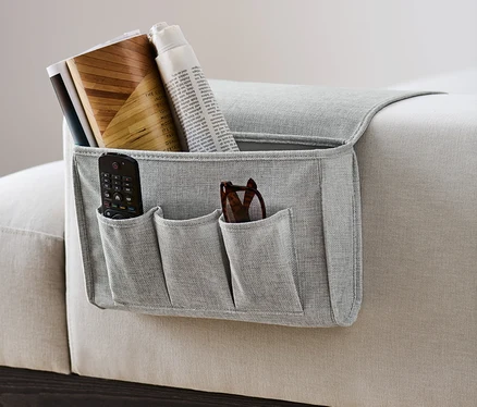 Grauer Bett-und-Couchorganizer hängt an der Seite eines Sofas und enthält eine Fernbedienung, eine Brille, Bücher und eine Zeitung.