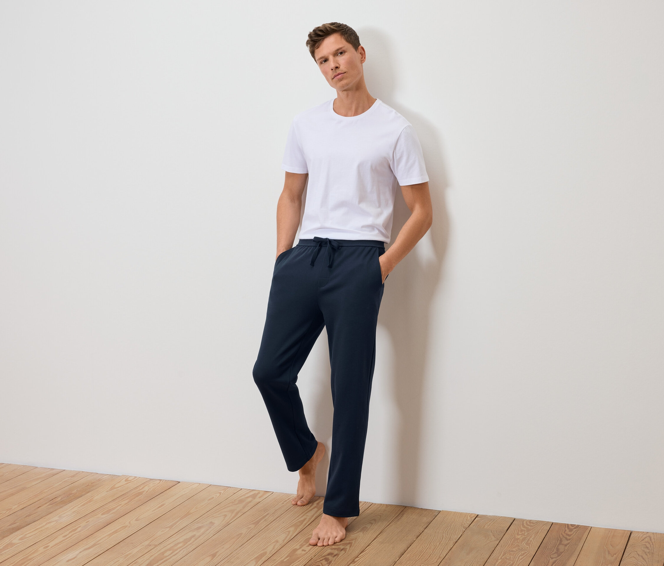 Mann steht an einer Wand gelehnt in einem weißen T-Shirt und dunkelblauen Jogginghose.