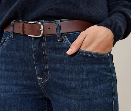 Detailaufnahme von Jeans mit einem cognacfarbenen Ledergürtel, eine Hand steckt in der Hosentasche.