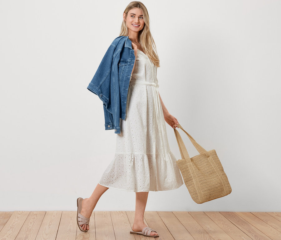 Eine Frau geht mit einer Jeansjacke über der Schulter, einem Shopper in Bastoptik und einem Blusenkleid mit Lochstickerei.